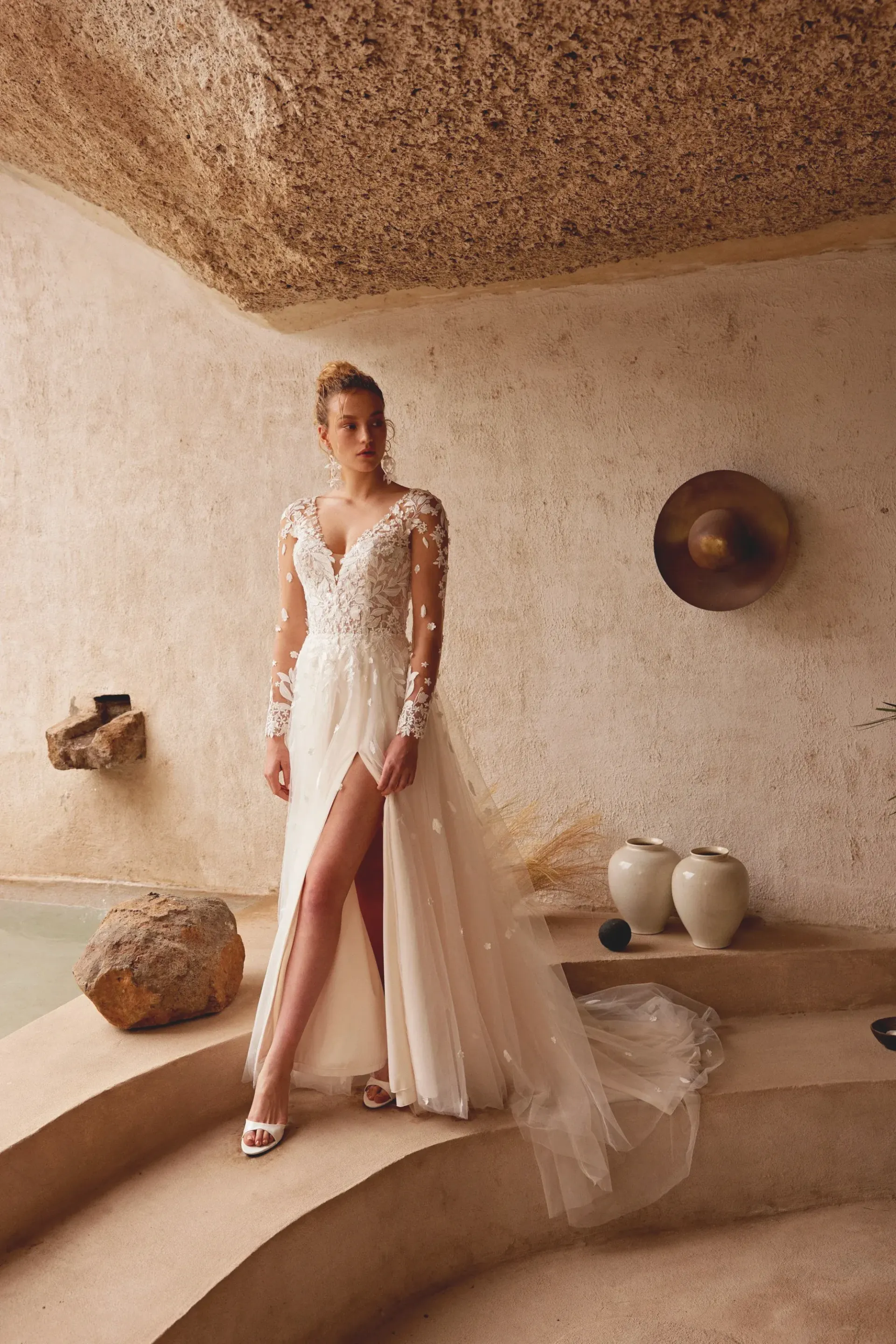 Woman in wedding dress with high slit, long sleeves, in stone-walled setting.