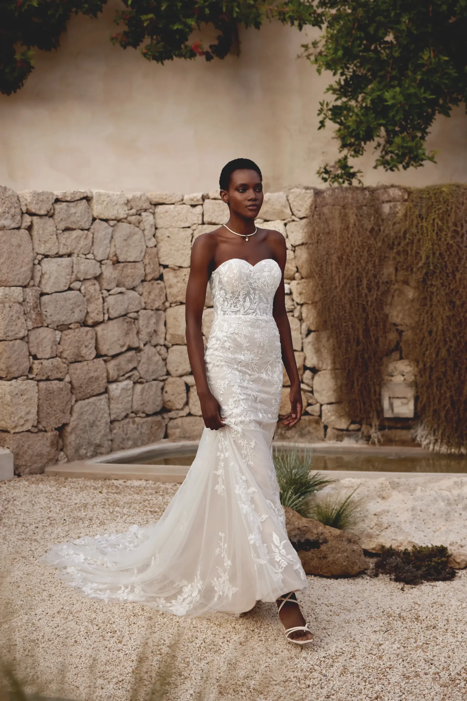 Woman in a strapless white lace wedding dress stands outside near a stone wall.