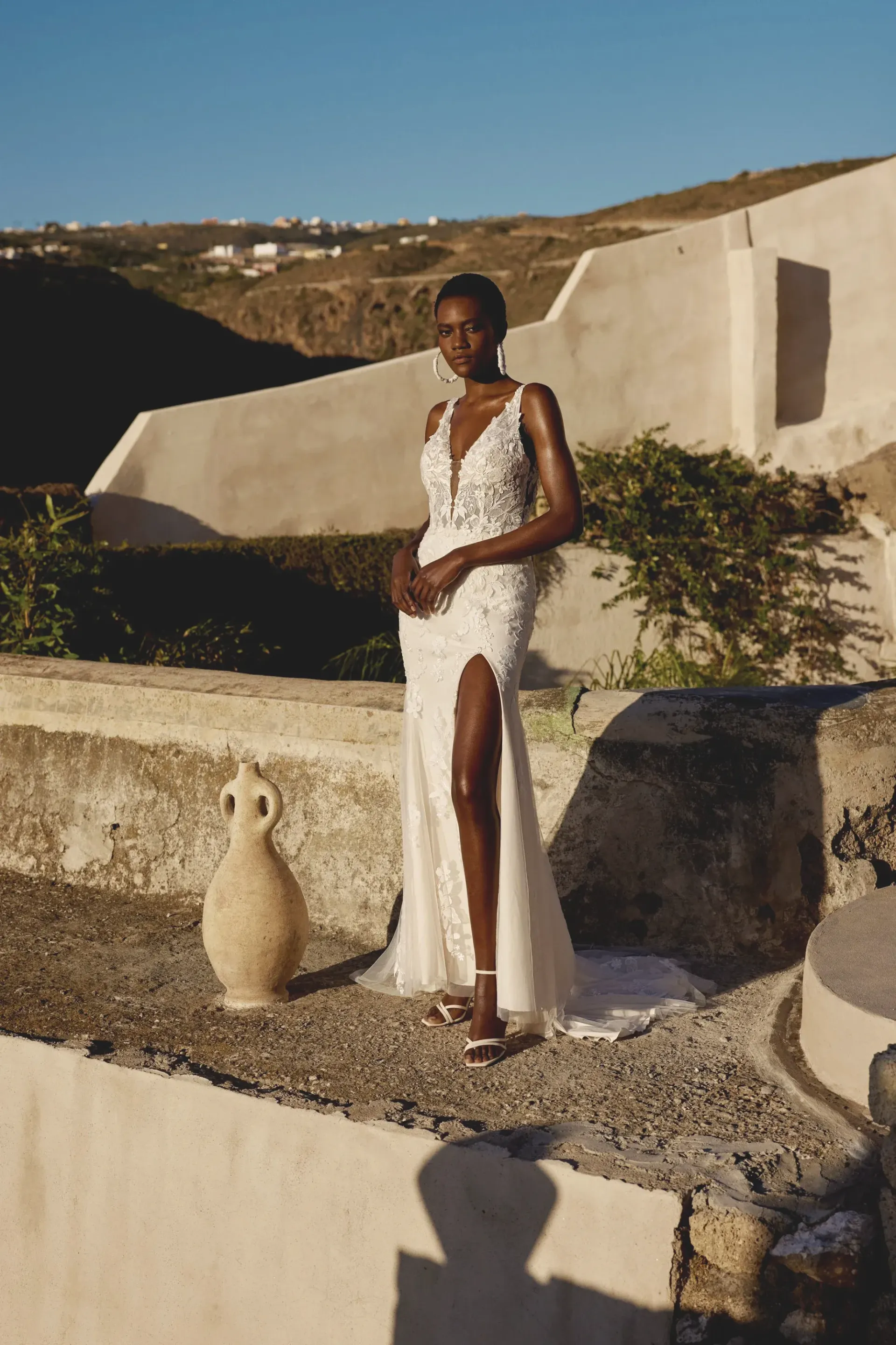 Woman in white beaded gown, high slit, poses outdoors. Santorini background, bright sunlight.
