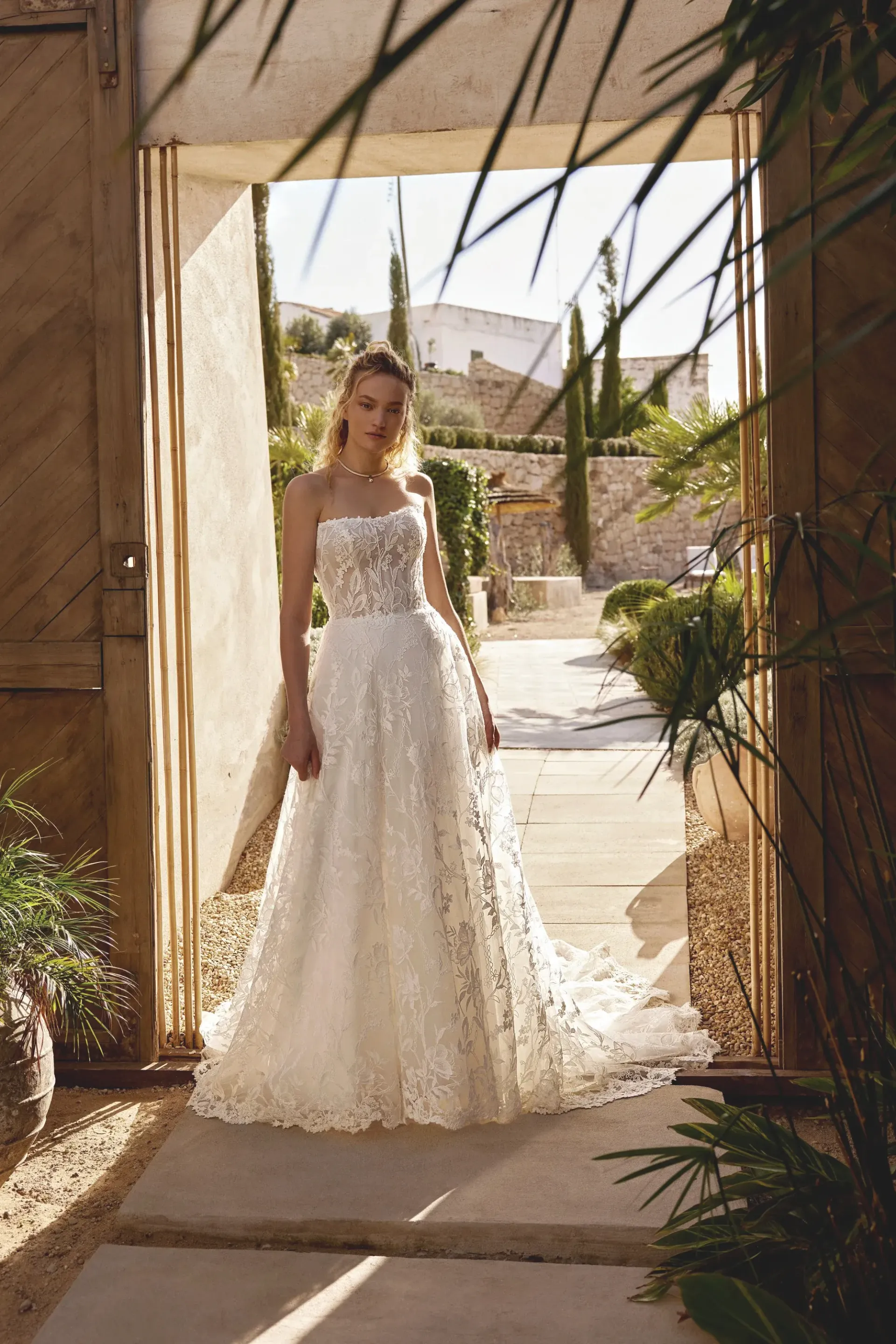 Woman in a white lace wedding dress stands in a stone doorway, looking out. Sunny outdoor scene.