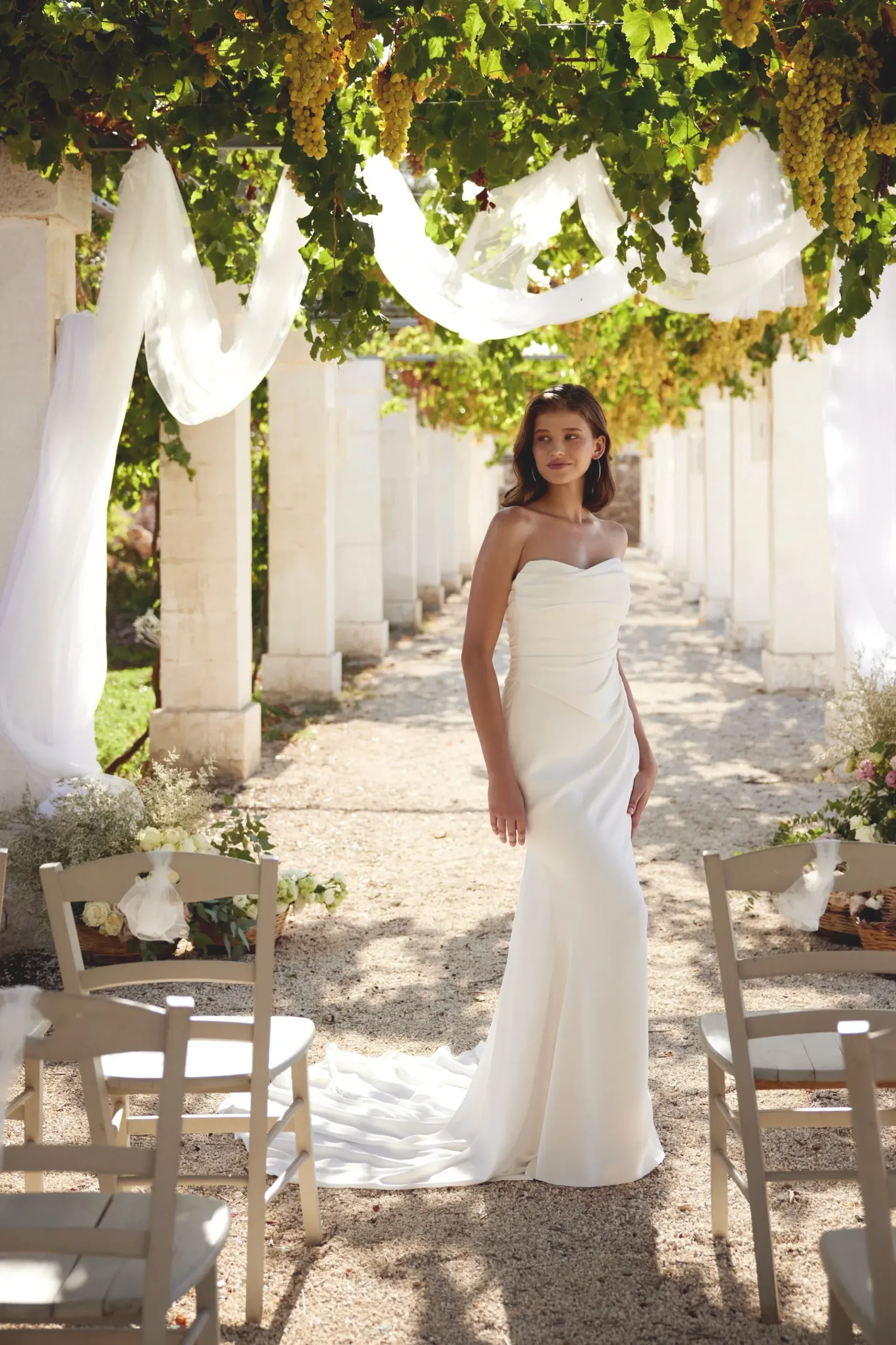 Bride in a white gown stands under a vine-covered pergola, ready for a wedding ceremony.