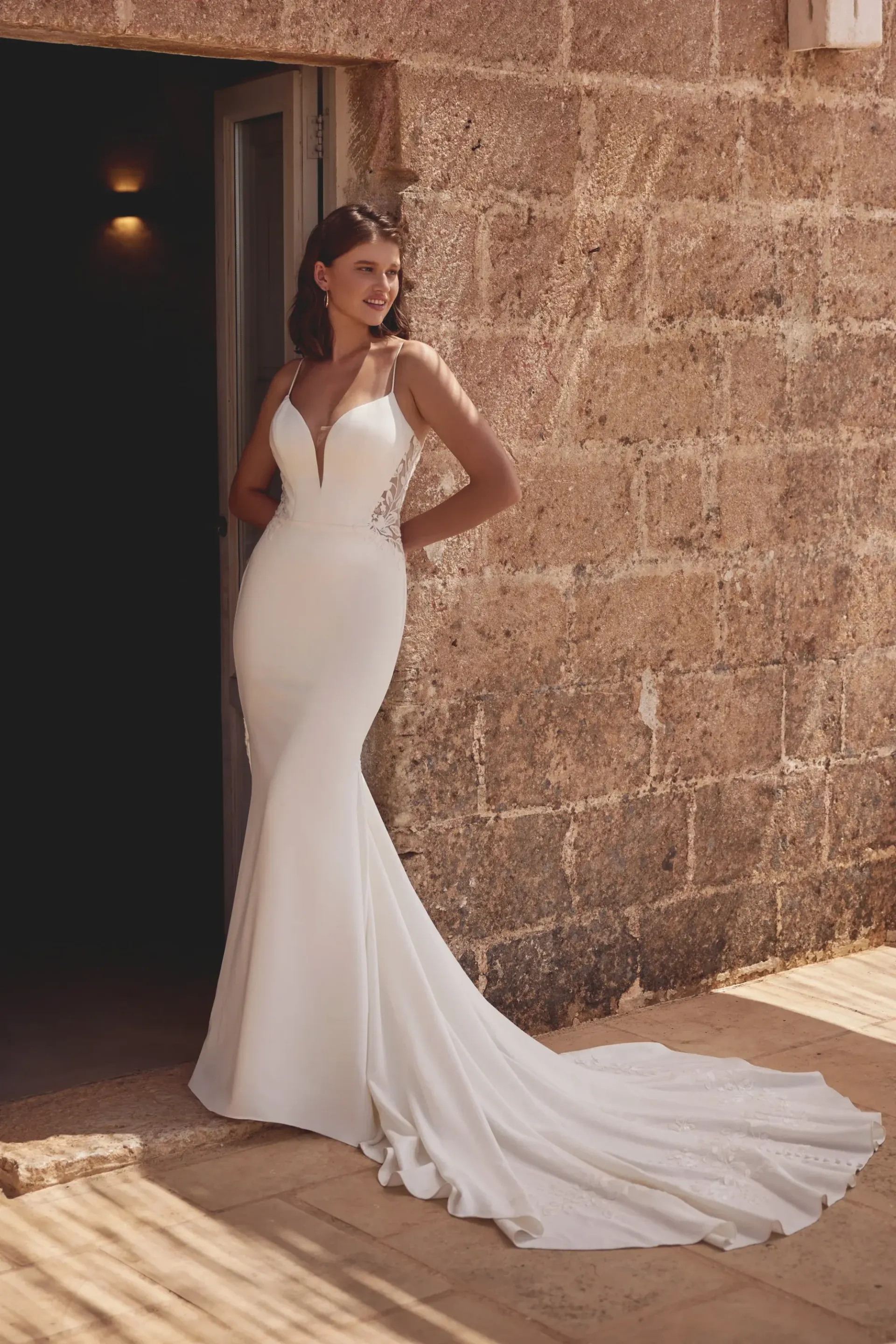 Woman in a white wedding dress with a long train poses near a stone wall.