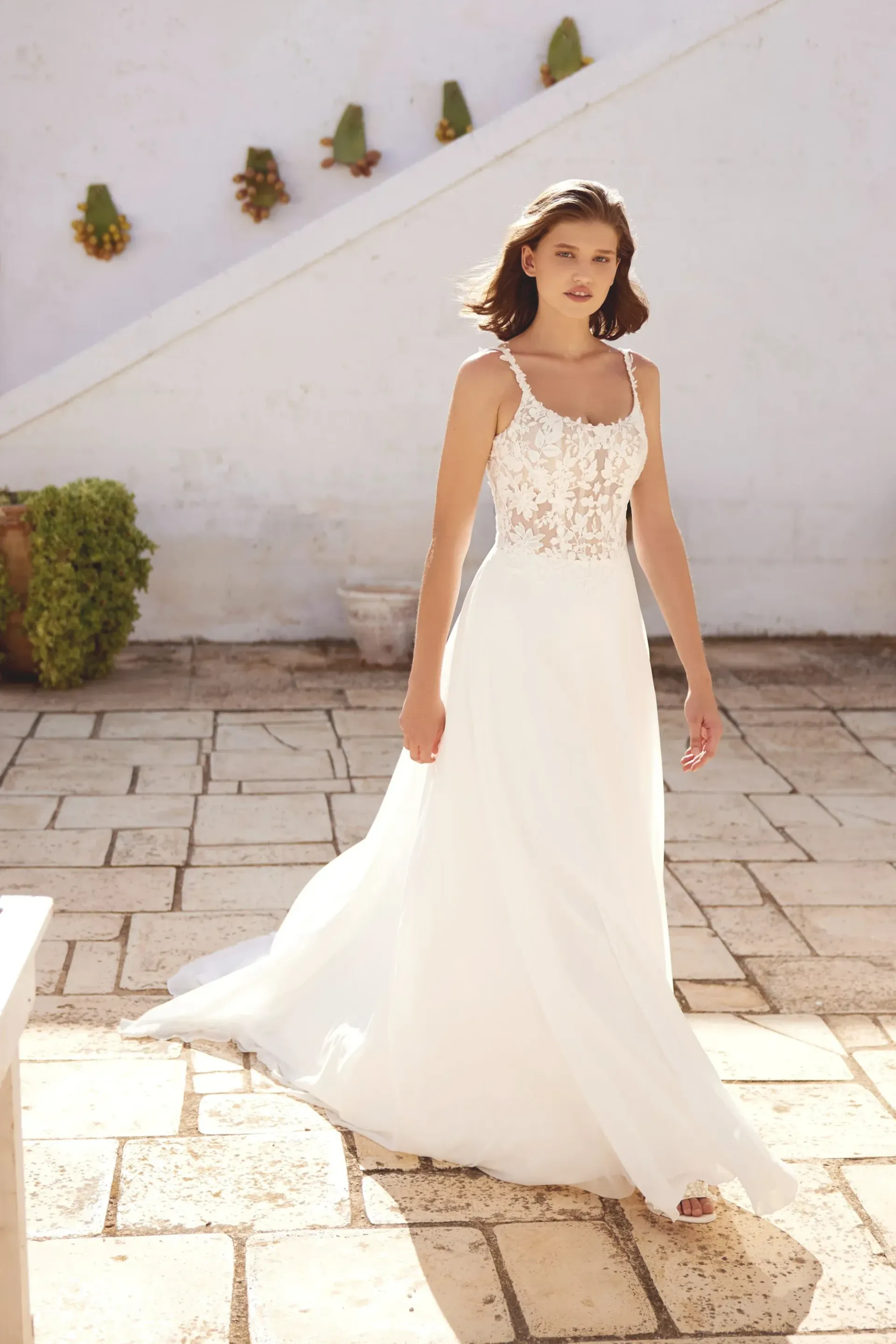 Woman in wedding dress, walking towards camera; outdoor stone patio, white building background.
