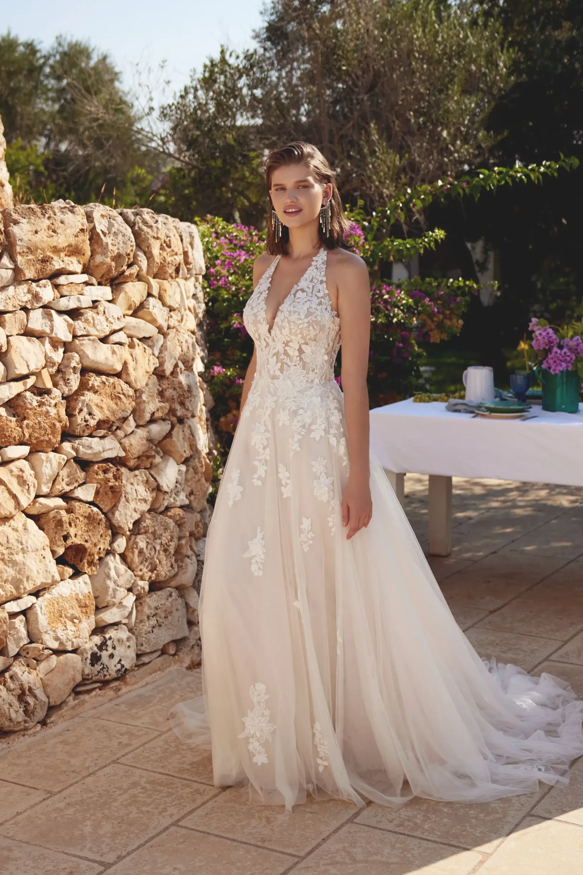 Woman in a wedding dress stands by a stone wall outdoors, with a table set for a celebration.