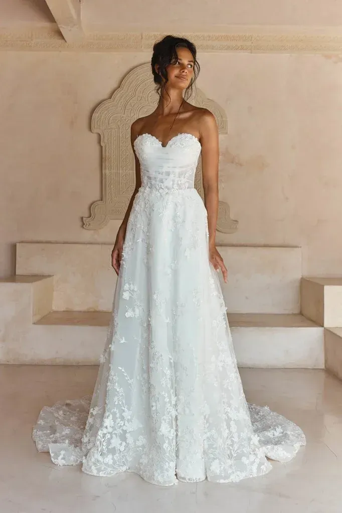 Woman in a white strapless wedding dress with floral detailing, smiling in a light-filled space.