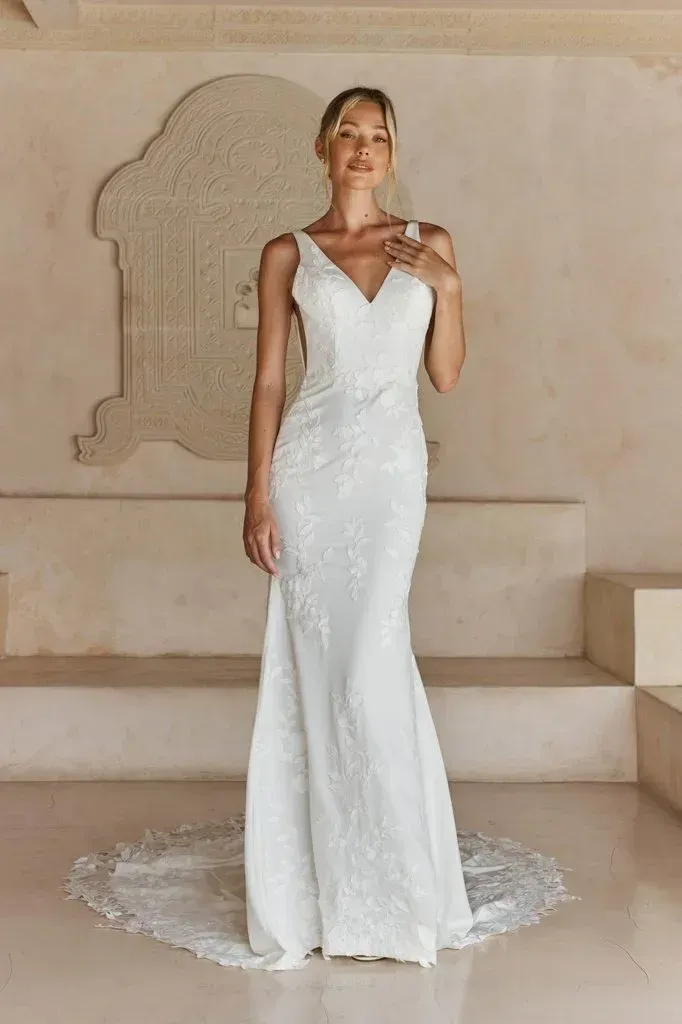 Woman in a white wedding dress stands on steps, smiling, inside a building with an ornate wall design.