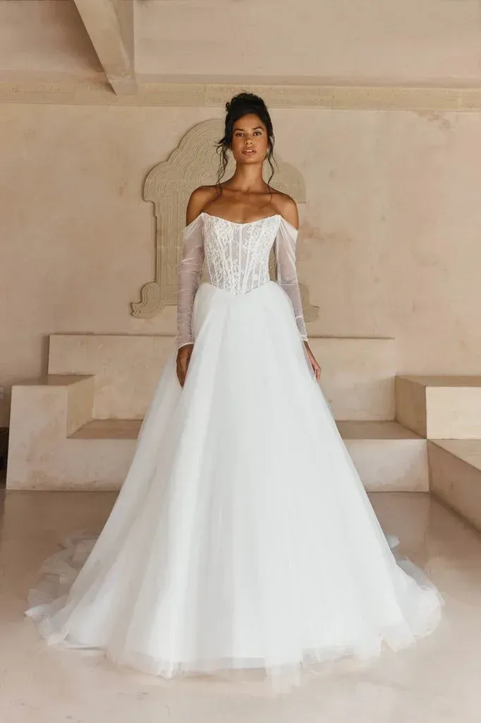 Woman in white off-the-shoulder wedding dress, long sleeves, standing in a light-filled room.