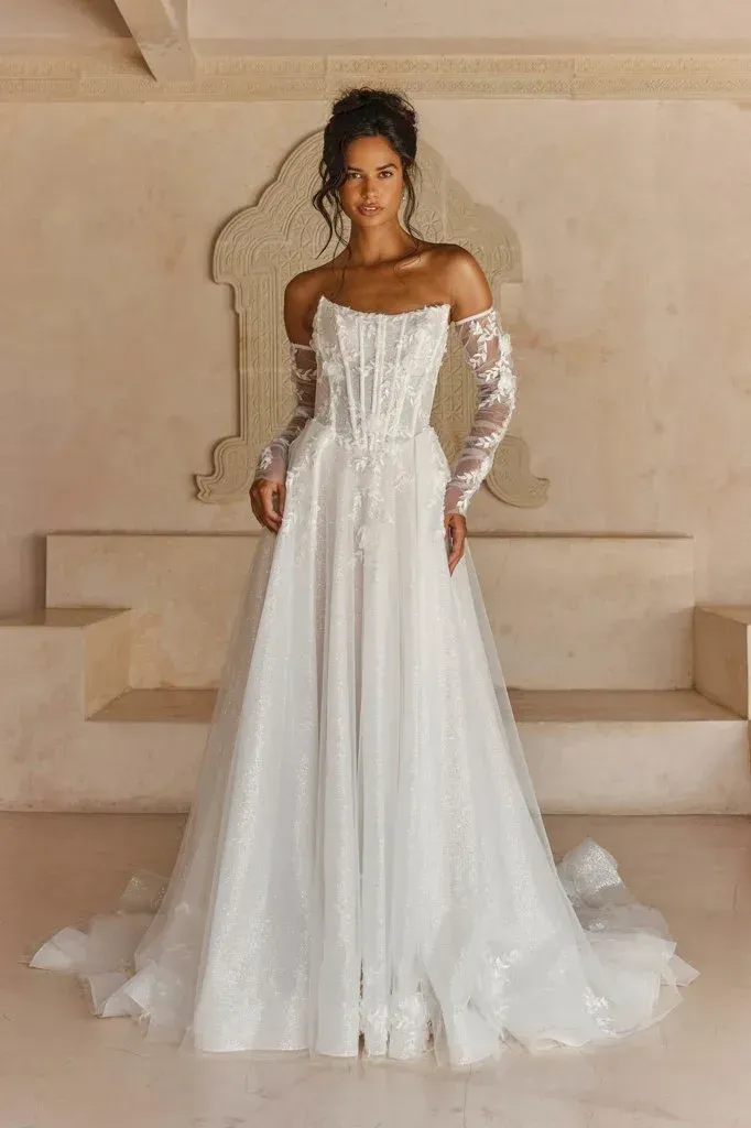 Woman in white off-the-shoulder wedding dress with lace sleeves, posed in an architectural setting.