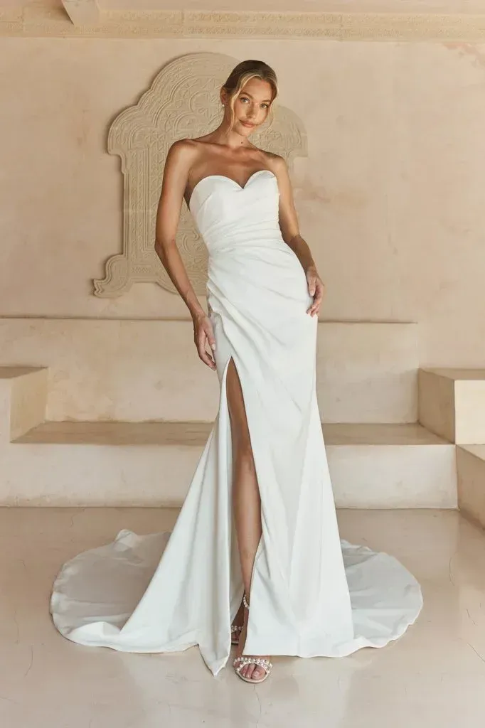 Woman in white strapless wedding dress with thigh-high slit, posing indoors.
