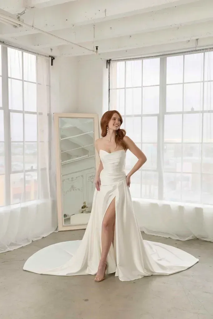 Woman in a white strapless wedding dress with a high slit, standing by a window; smiling.