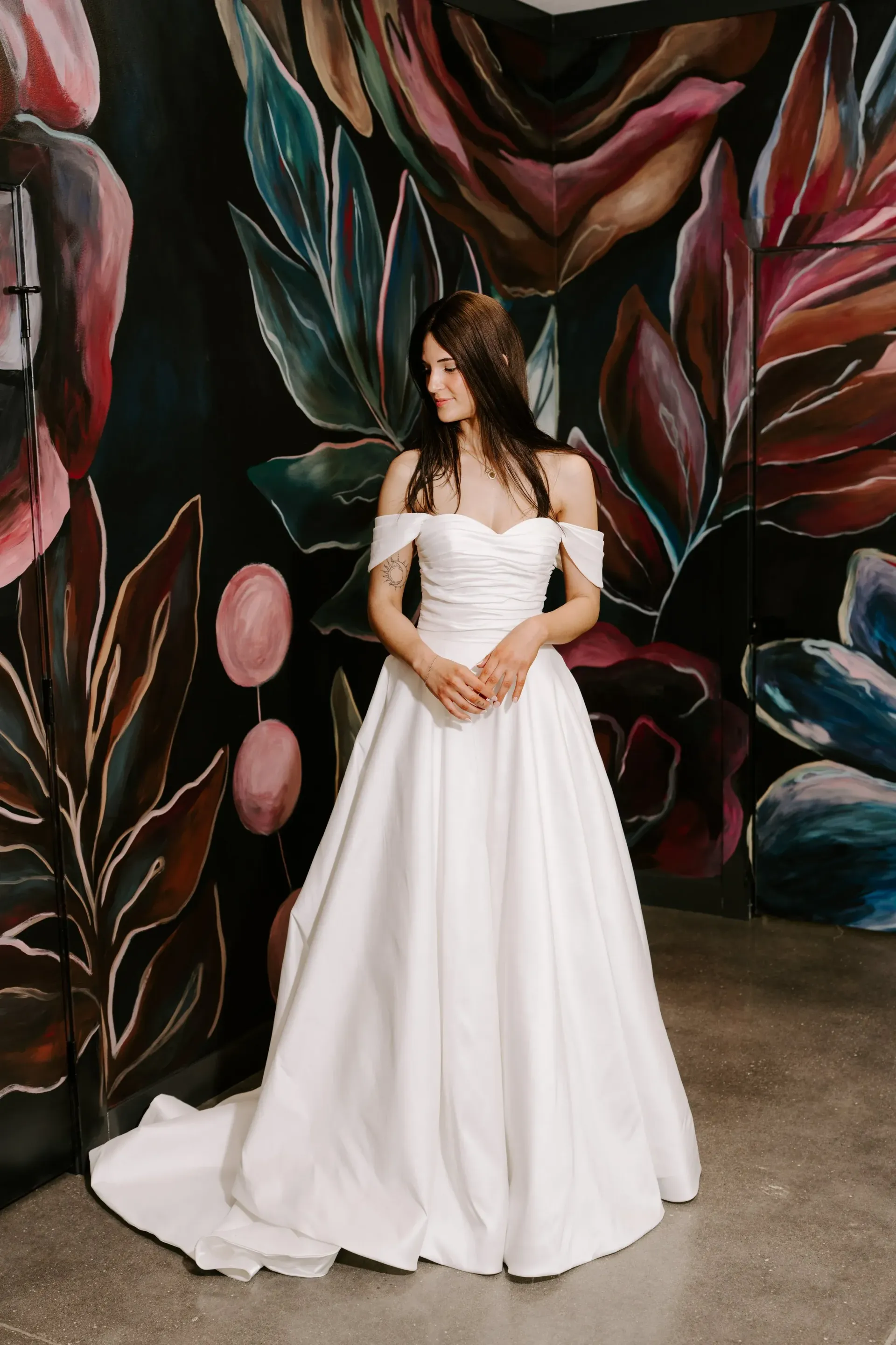 Woman in white off-the-shoulder wedding dress stands before a colorful floral mural, smiling softly.