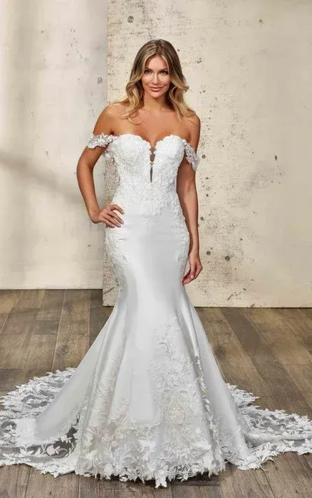 Woman in a white off-the-shoulder wedding dress with lace details and train, standing in a studio.