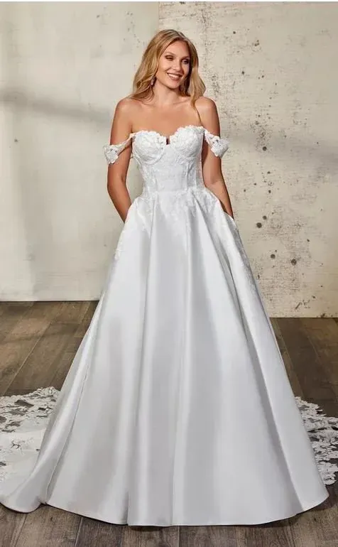 Blonde woman in white off-the-shoulder wedding dress with pockets, smiling. Rustic background.