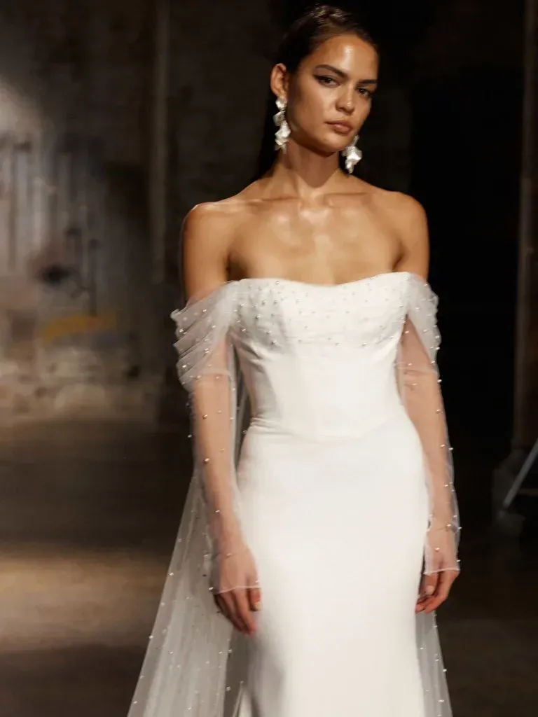 Woman in a white strapless wedding dress with sheer pearl-adorned sleeves and train, standing on a runway.