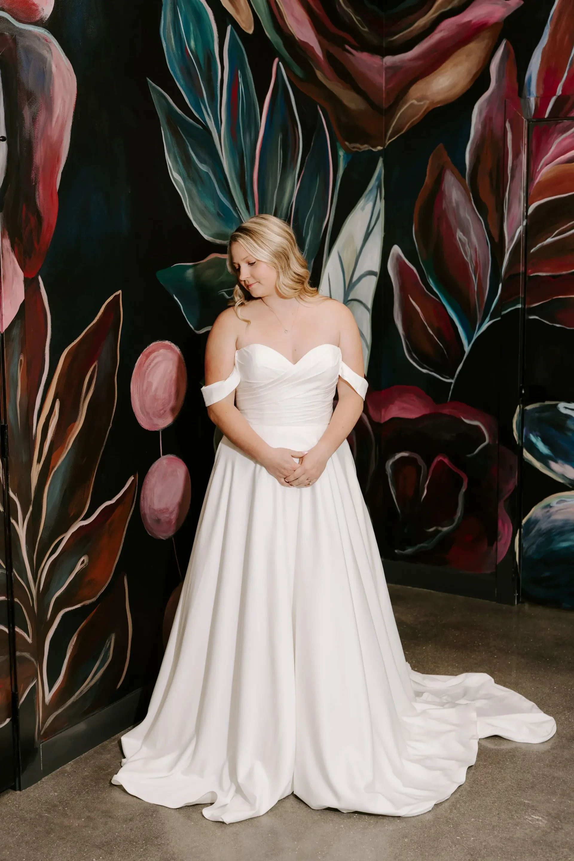 Woman in white wedding dress, off-shoulder neckline, hands clasped, stands before floral mural.