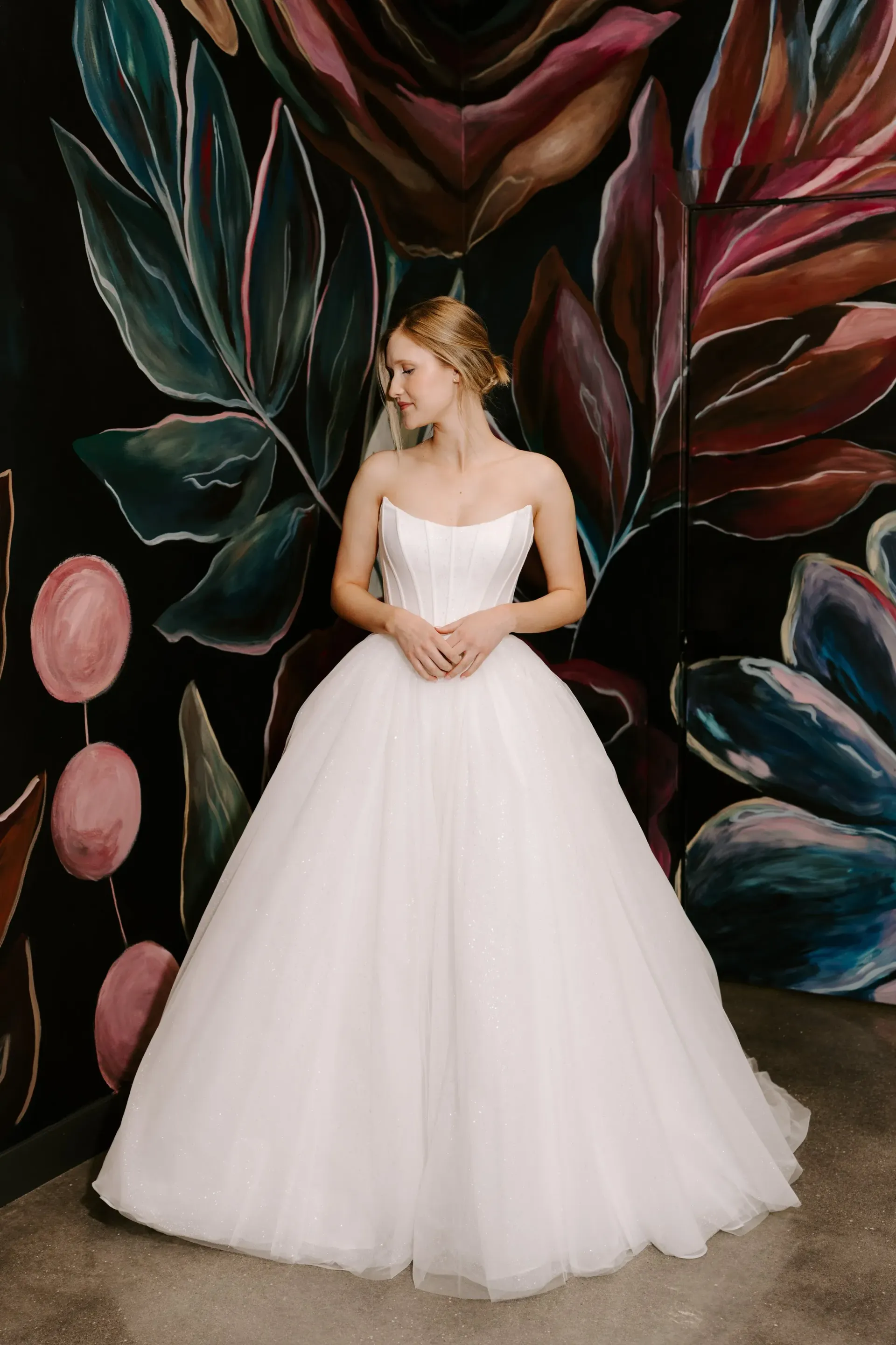 Blonde bride in white ballgown poses against a floral painted wall, smiling.