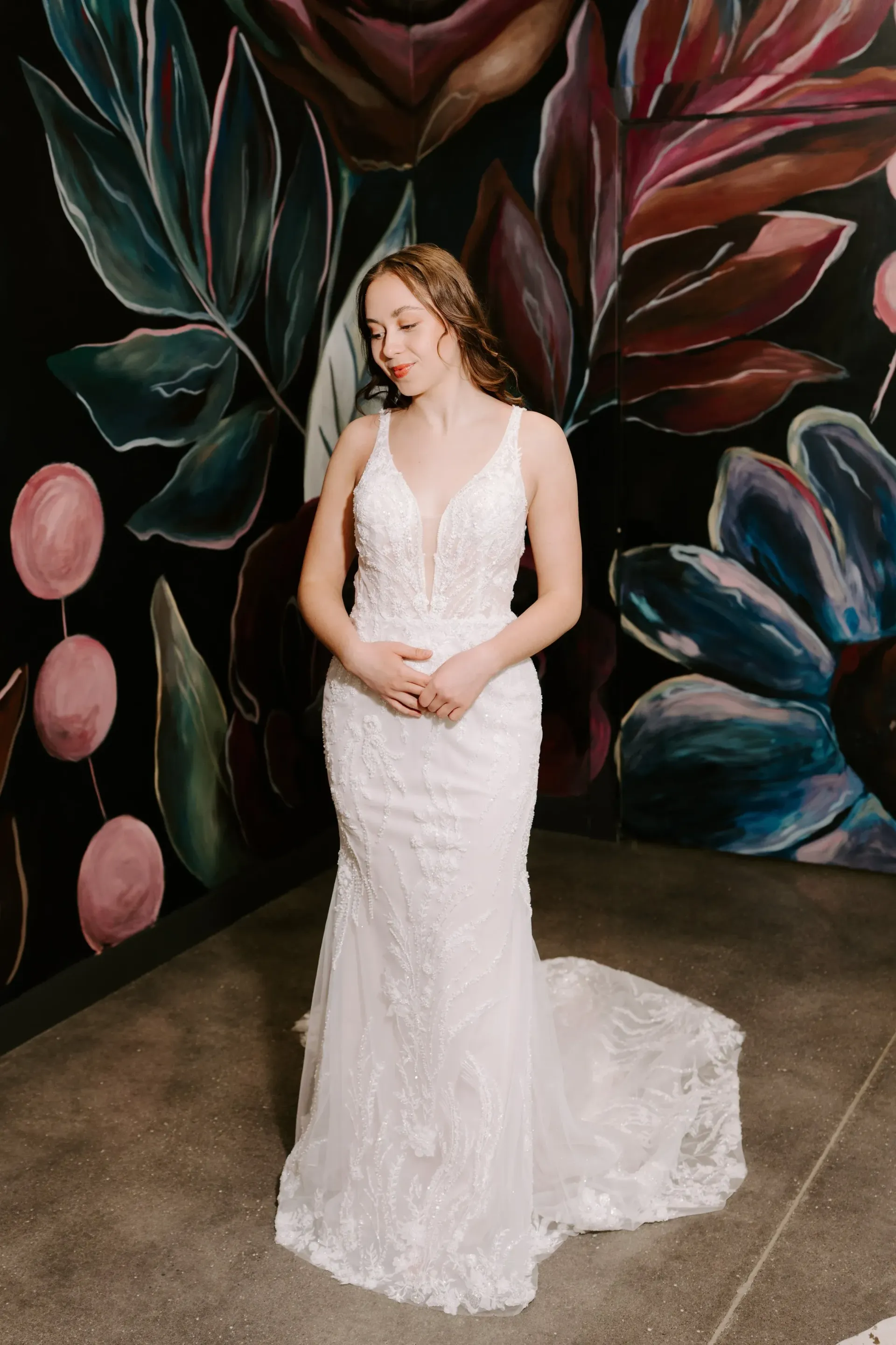 Bride in white lace gown stands before a black wall with floral mural.