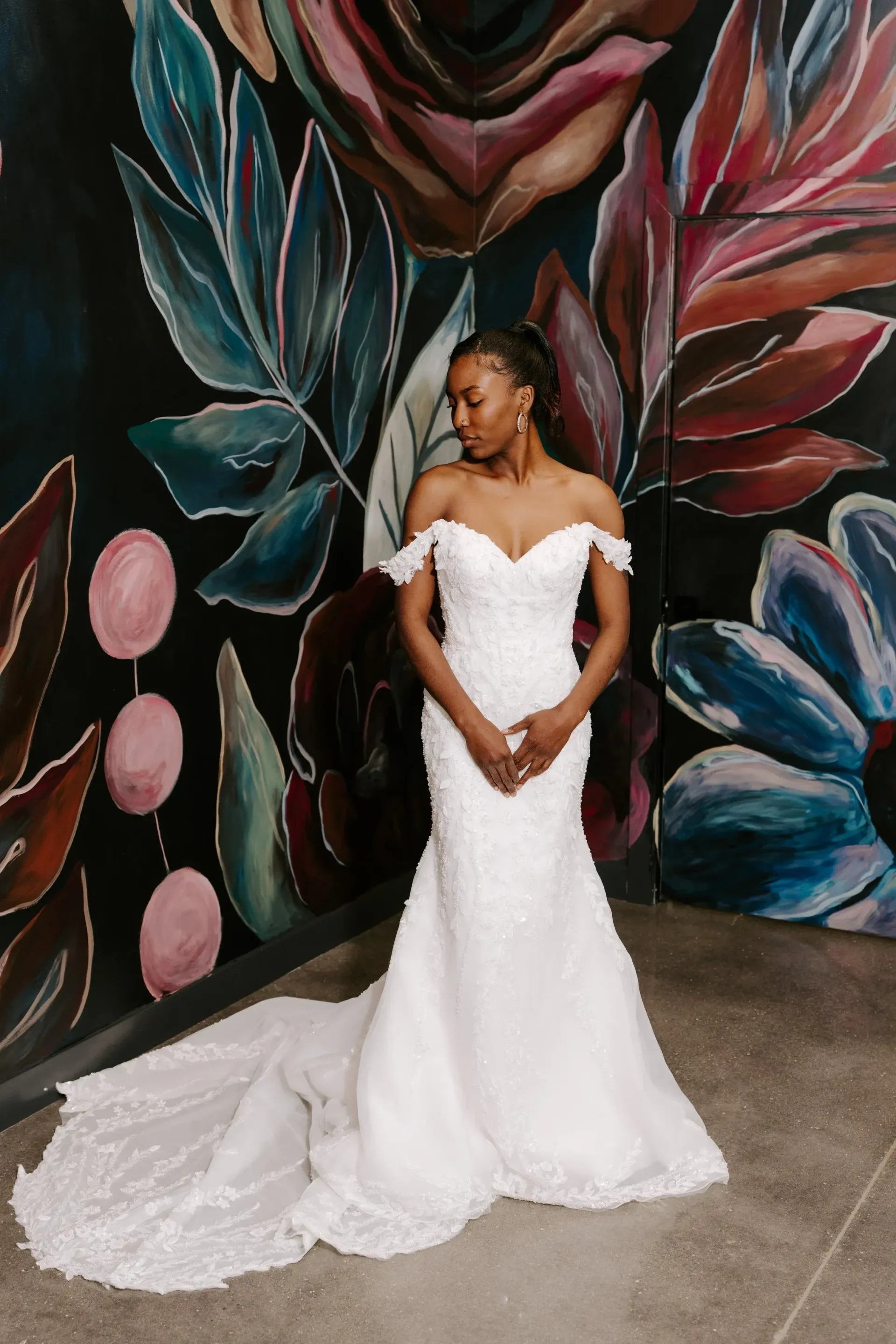 Bride in white off-shoulder gown poses in front of a floral mural.