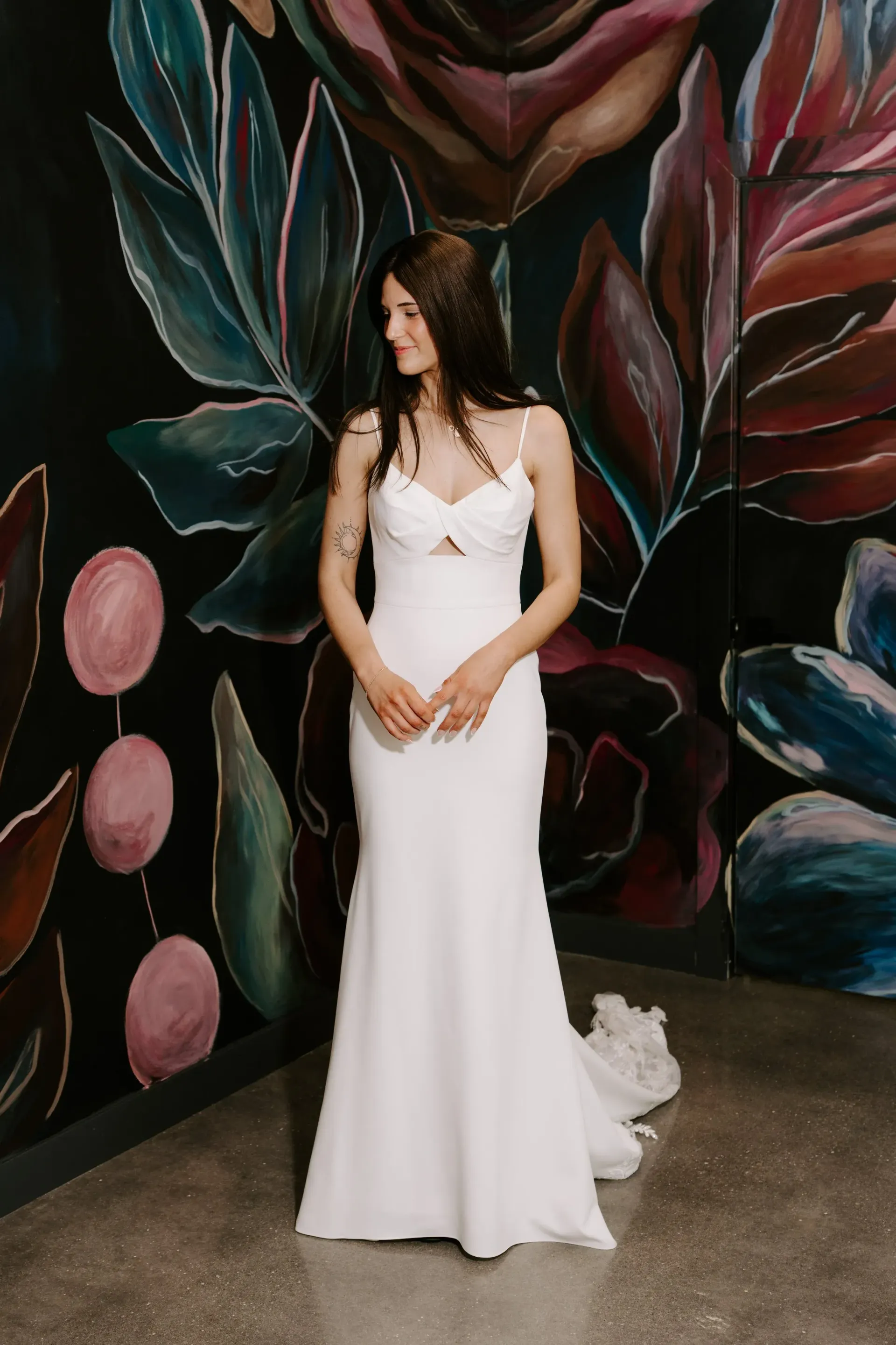 Woman in white dress stands by floral mural, smiling.
