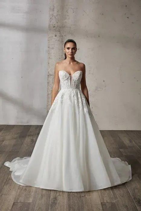 Woman in a white strapless wedding gown with a long train, standing in a neutral-toned room.