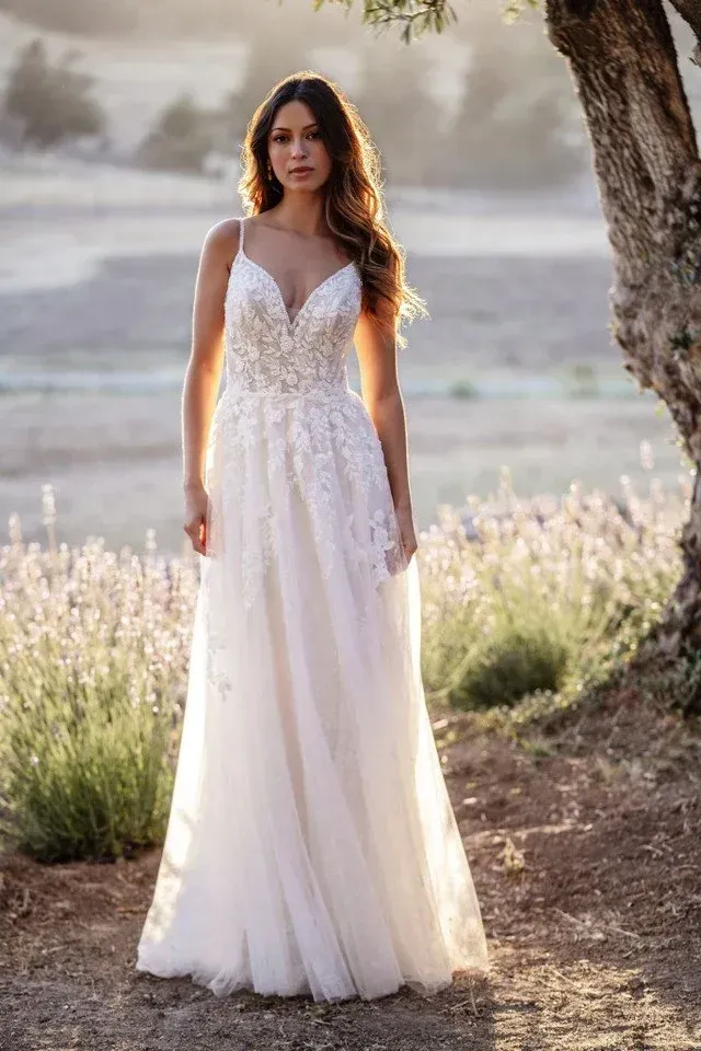Woman in a white wedding dress poses outdoors; field and tree in background, sunlight.