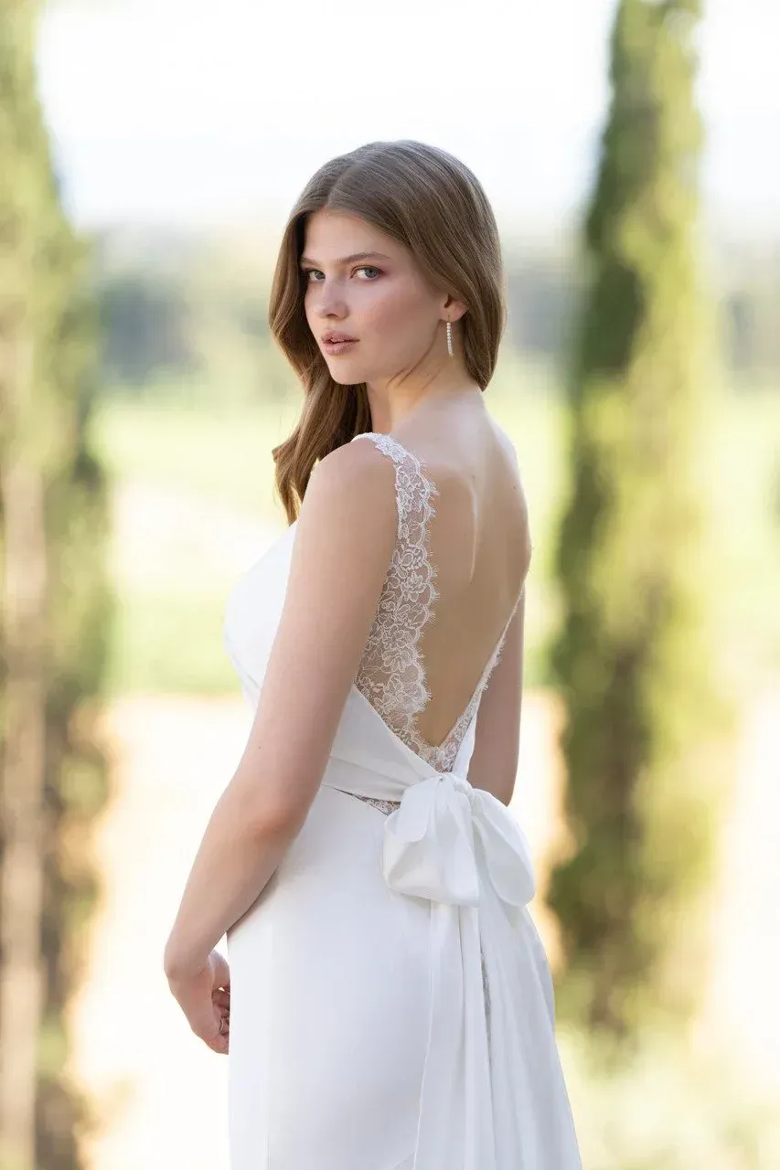 Woman in a white wedding dress with a low lace back and bow detail, looking over her shoulder outdoors.