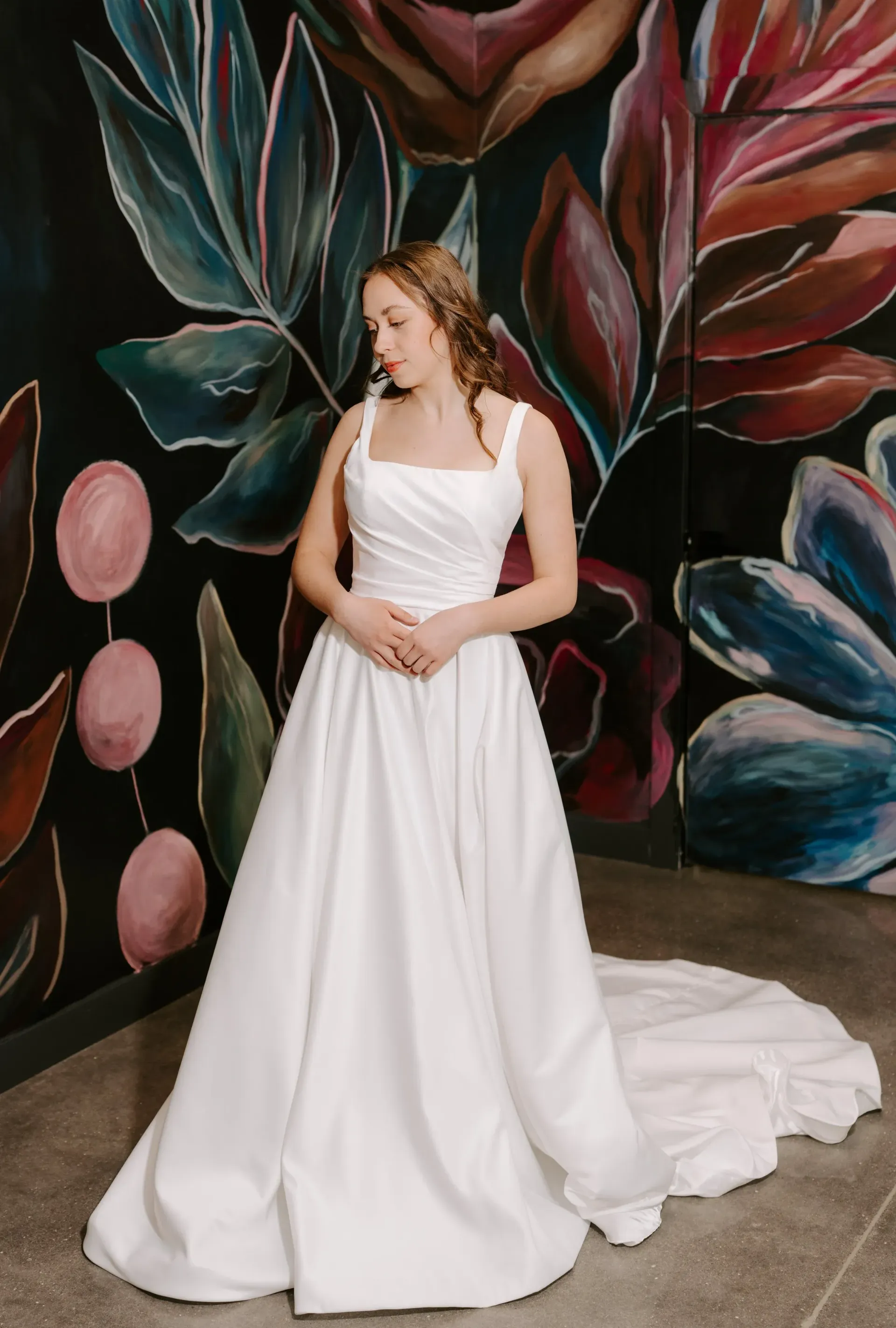 Woman in a white wedding dress with a long train, standing in front of a floral mural.