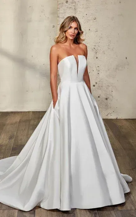 Woman in a strapless white wedding dress with a plunging neckline and pockets, standing in front of a textured wall.