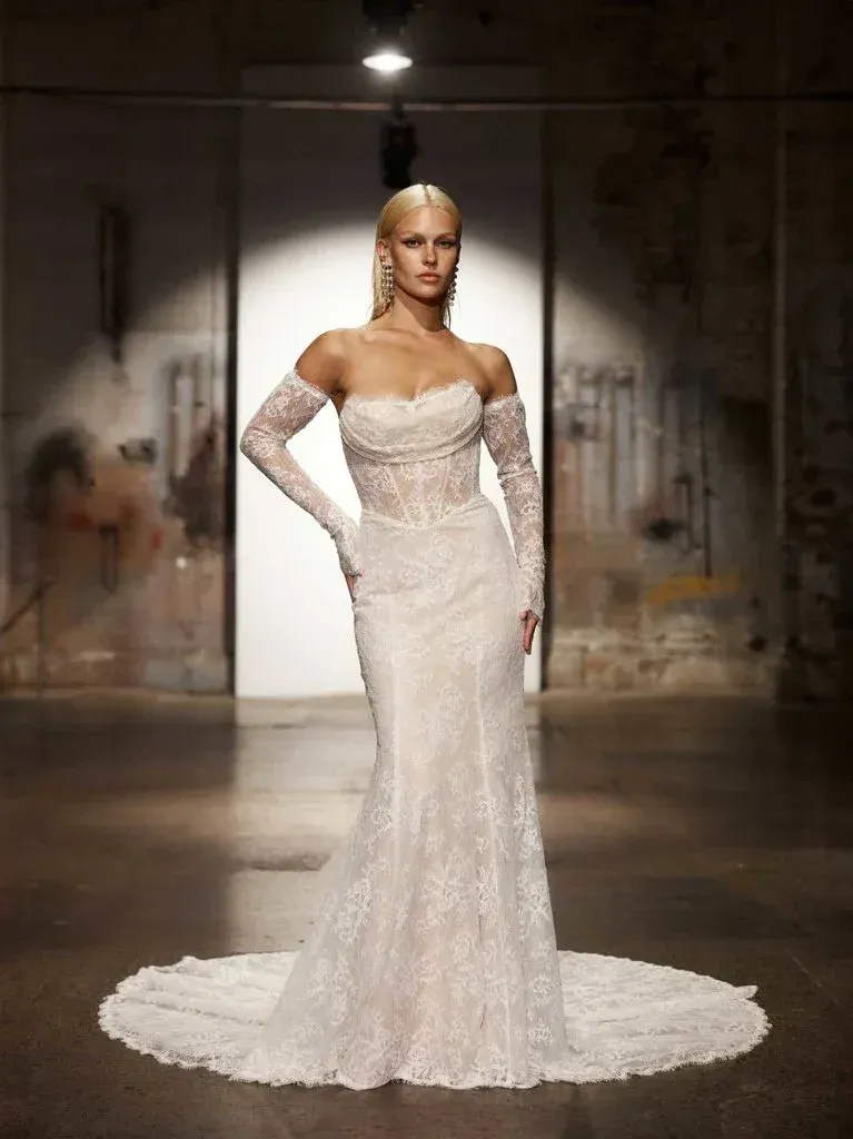 Woman in a white, lace mermaid-style wedding gown with off-the-shoulder gloves. Runway setting, blonde hair.