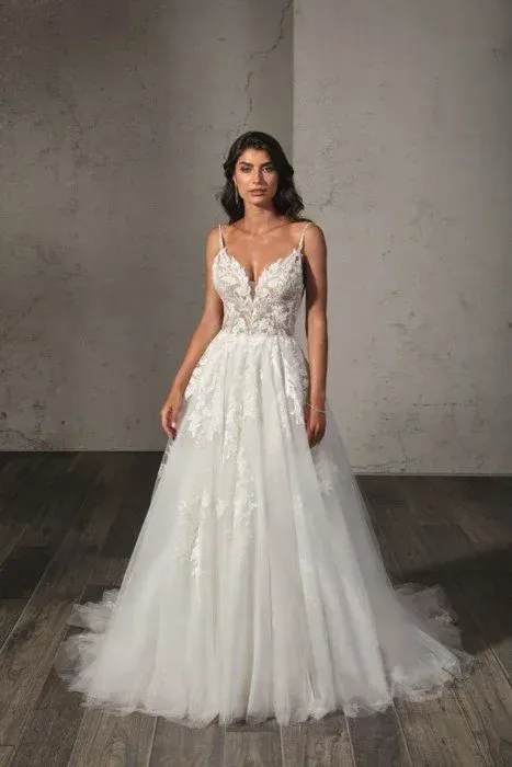 Woman in a white wedding dress with floral bodice, spaghetti straps, and a full skirt, standing indoors.