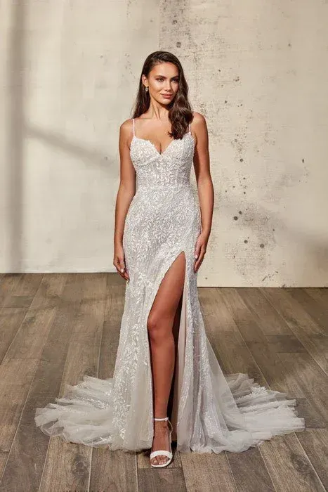 Woman in a white, beaded wedding dress with a thigh-high slit and open-toed heels, standing on a wood floor.