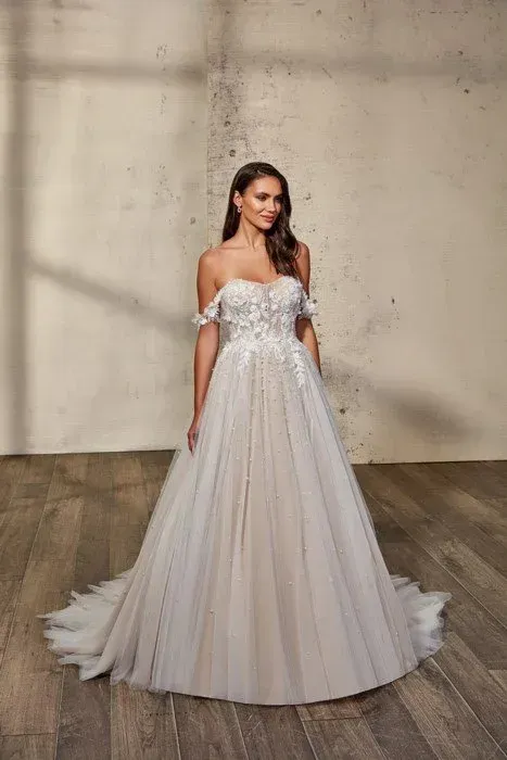 Woman in a white off-the-shoulder wedding dress, smiling, standing on a wooden floor.