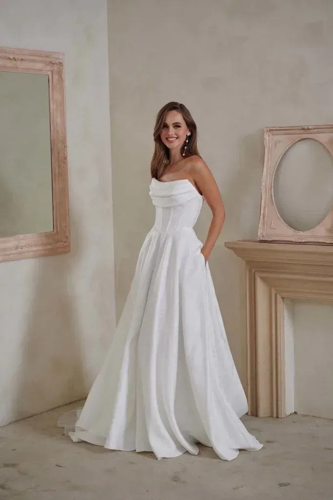 Woman in a strapless white wedding dress with pockets stands indoors, smiling.