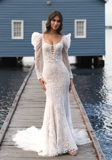 Woman in a white, lace wedding dress stands on a wooden dock, blue building in the background.
