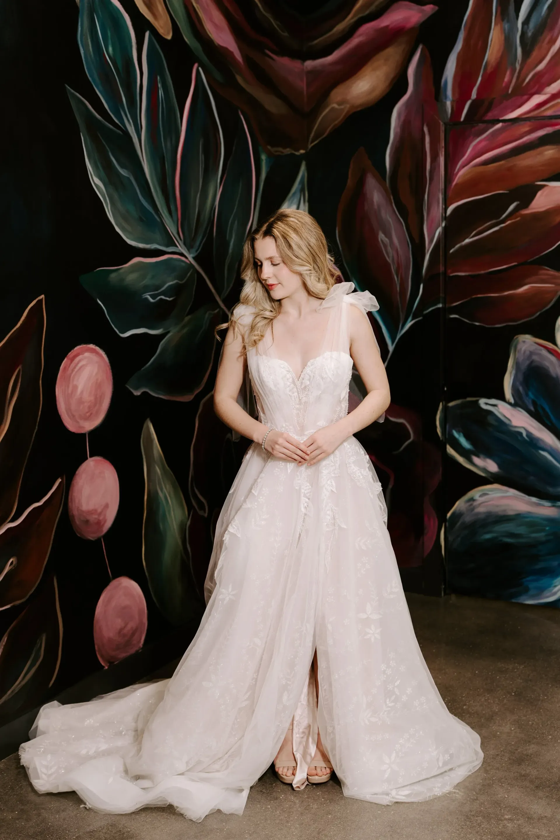 Blonde woman in a white wedding dress stands before a colorful floral mural, looking down.