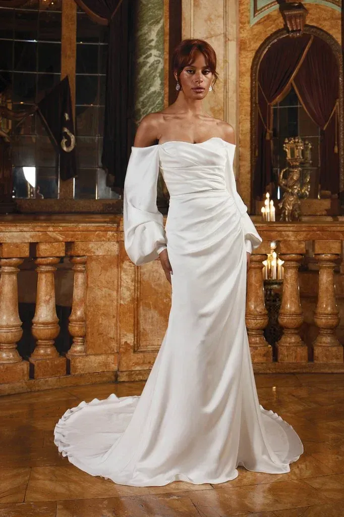 Woman in an off-the-shoulder white wedding dress with train, standing in ornate room.