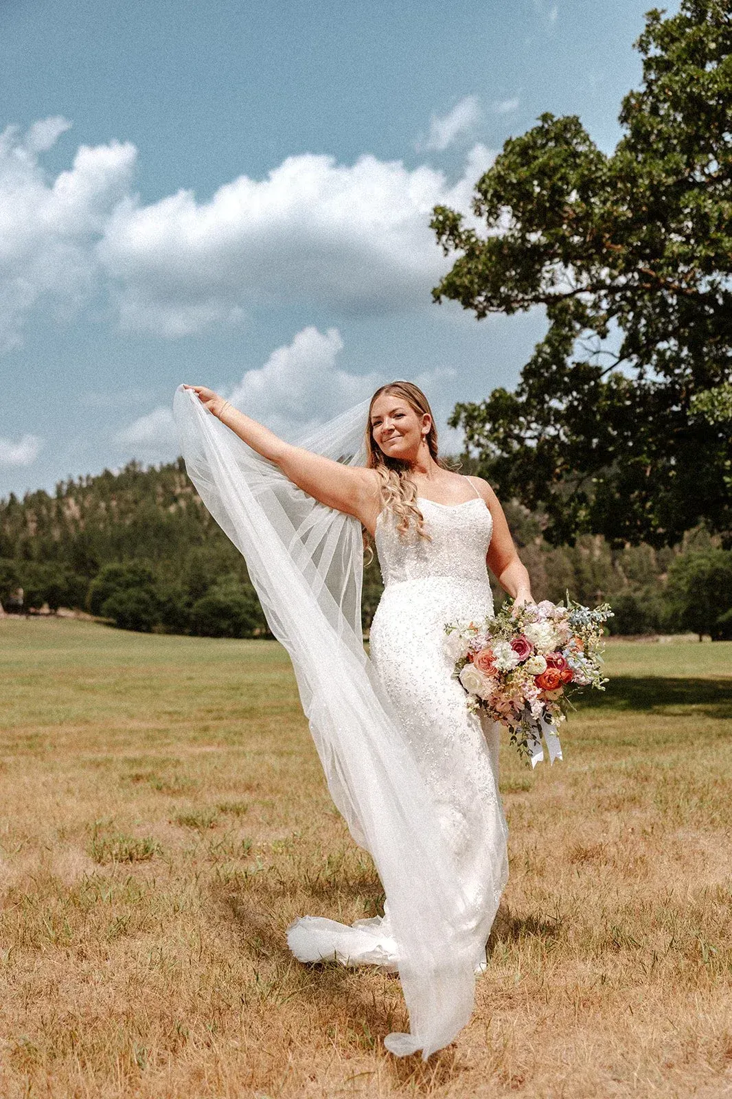 Bride in a white gown waving a veil, holding flowers, smiling, in a field.