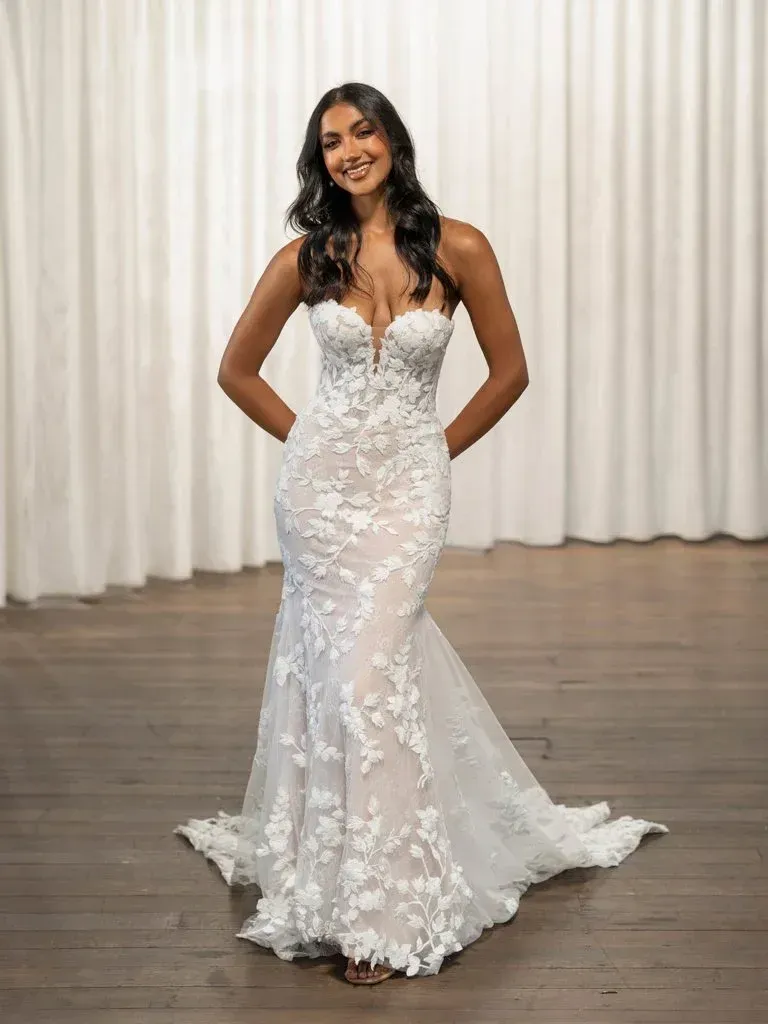 Woman in a white floral wedding gown smiles with hands behind her back.