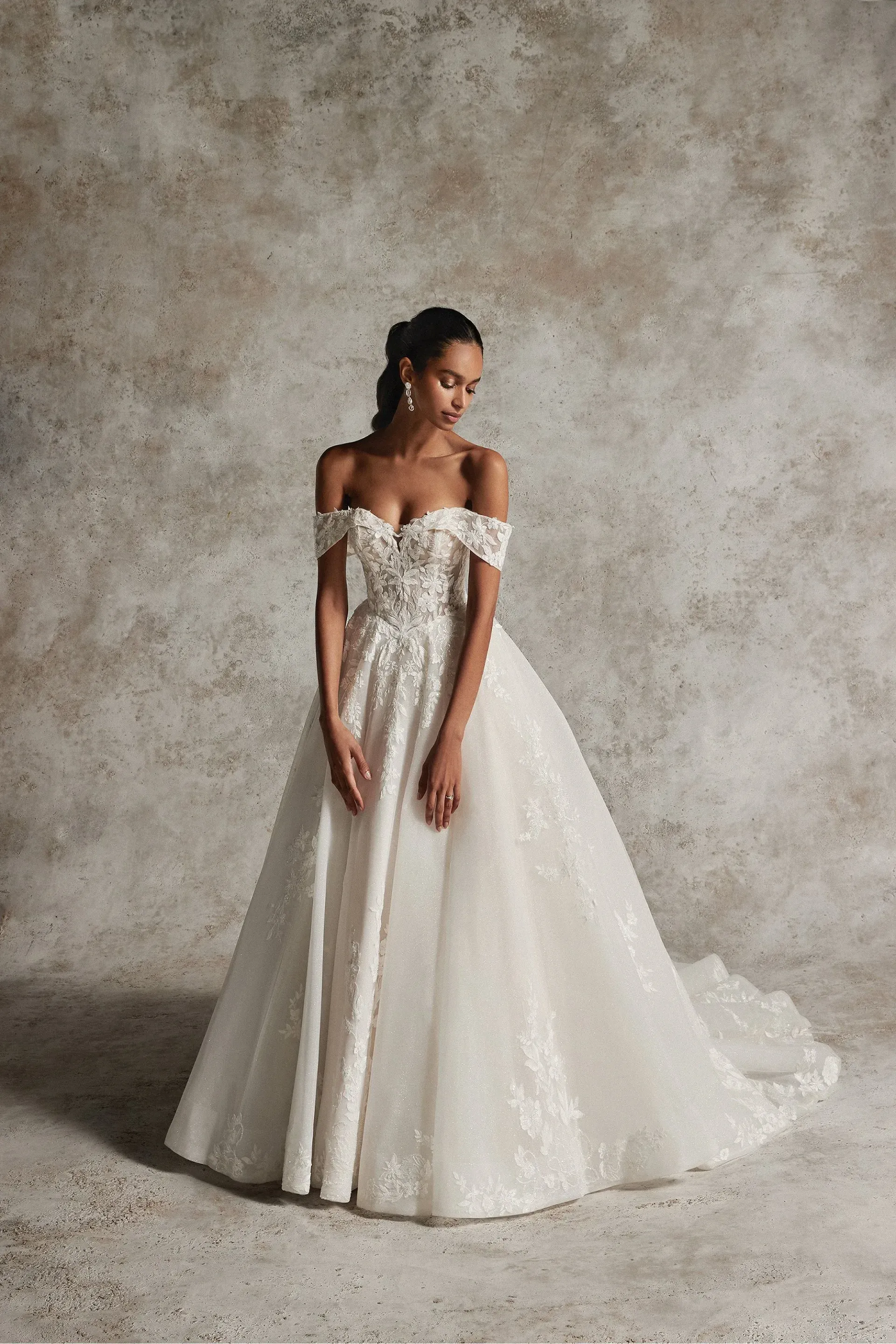 Woman in off-the-shoulder wedding dress, looking down, against a mottled neutral backdrop.