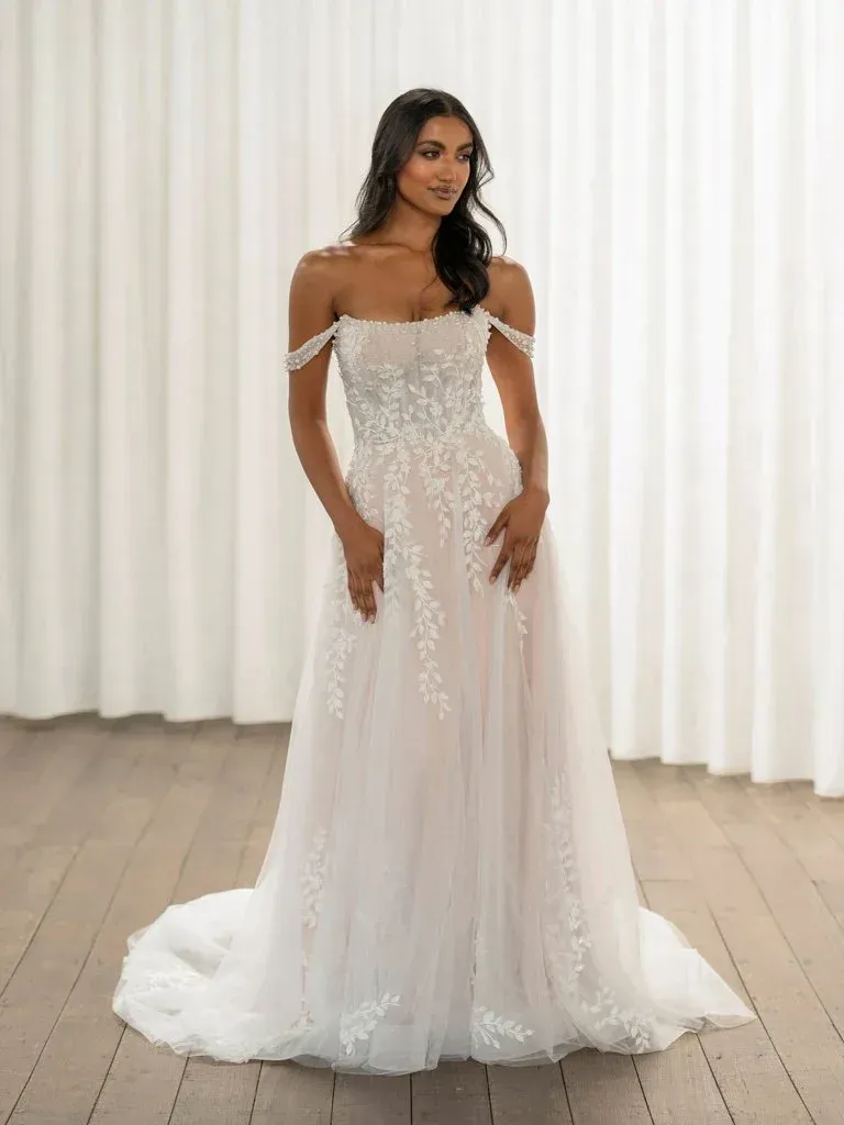 Woman in a white, off-the-shoulder wedding dress with lace appliques, standing in a room with white curtains.