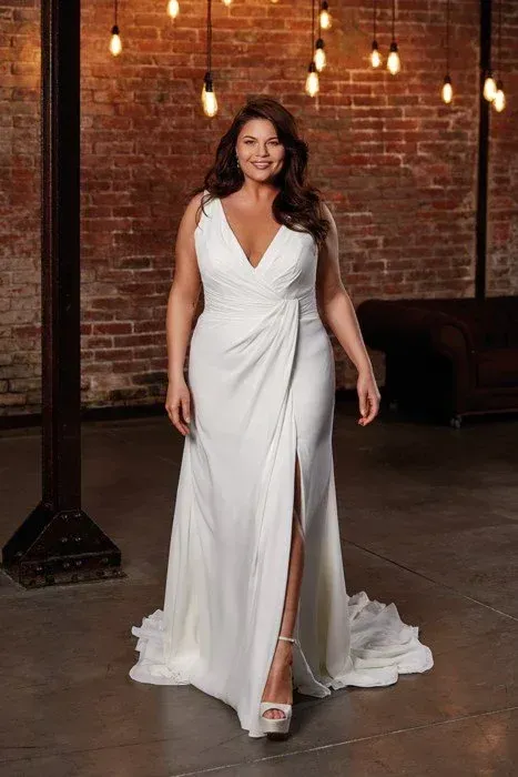 Woman in a white wedding dress with a thigh slit, smiling, standing in front of a brick wall.