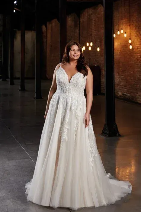 Woman in a white wedding dress with a beaded bodice, standing in a dark, industrial-looking space with brick walls.