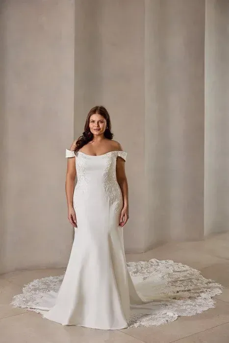 Woman in a white off-the-shoulder wedding dress with a long train, standing in a neutral-toned space.