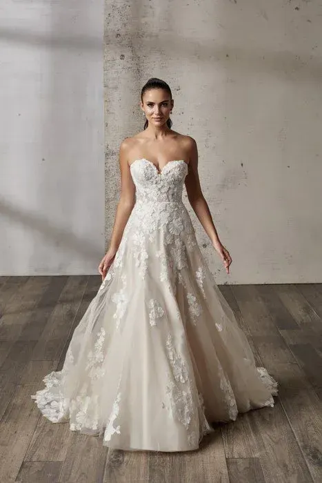 Woman in strapless white lace wedding dress, standing indoors with hands at sides, smiling.