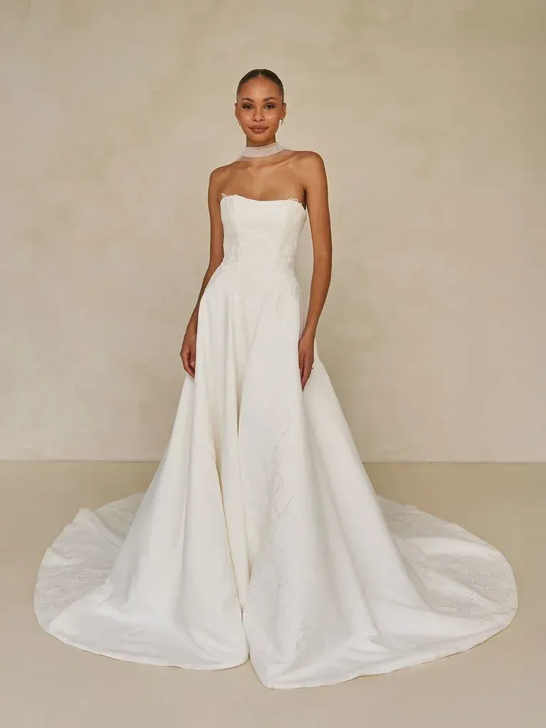 Woman in a strapless white wedding gown with a long train. Neutral background.