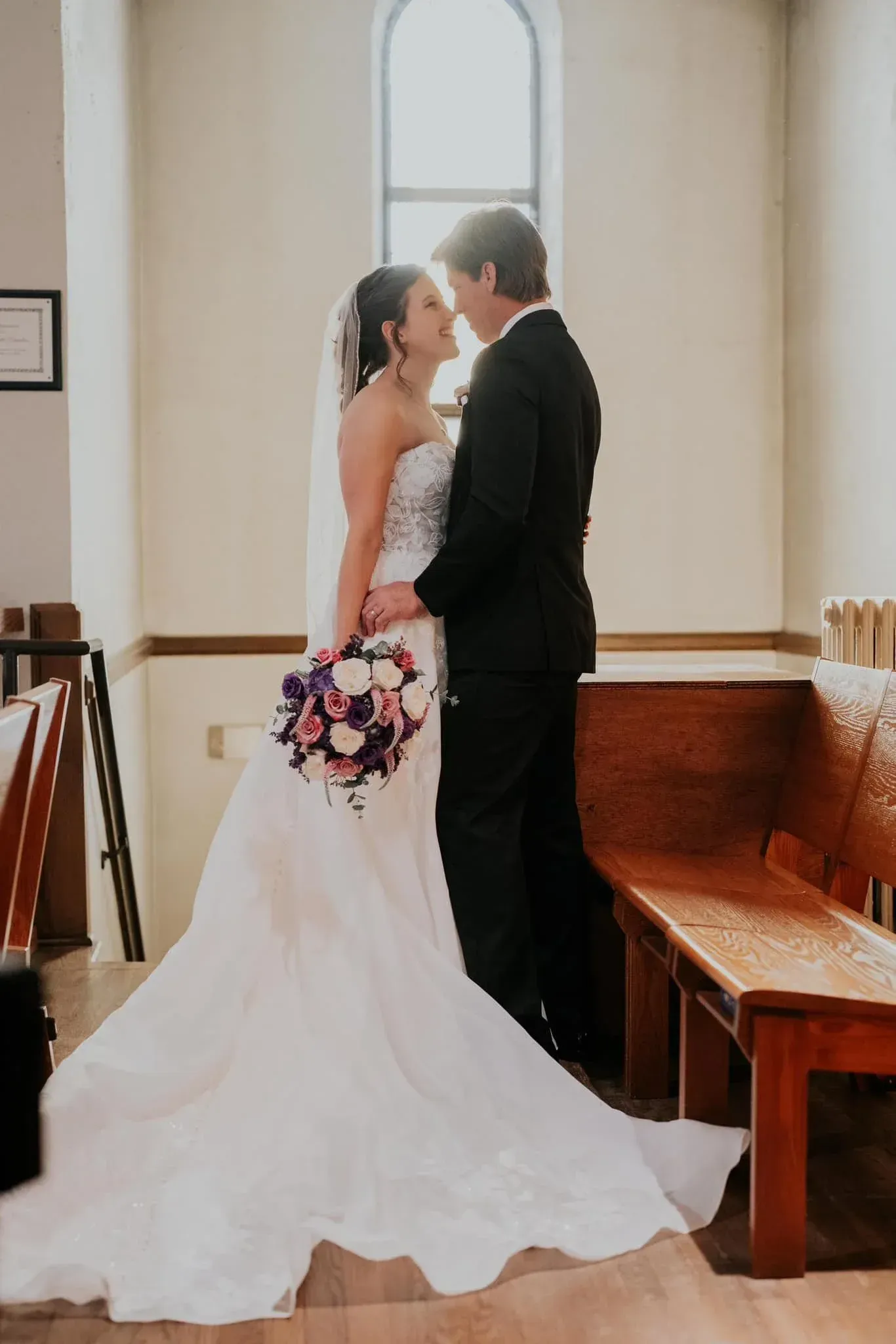 Bride and groom in a church, facing each other. Bride in white dress, holding bouquet. Groom in black suit.