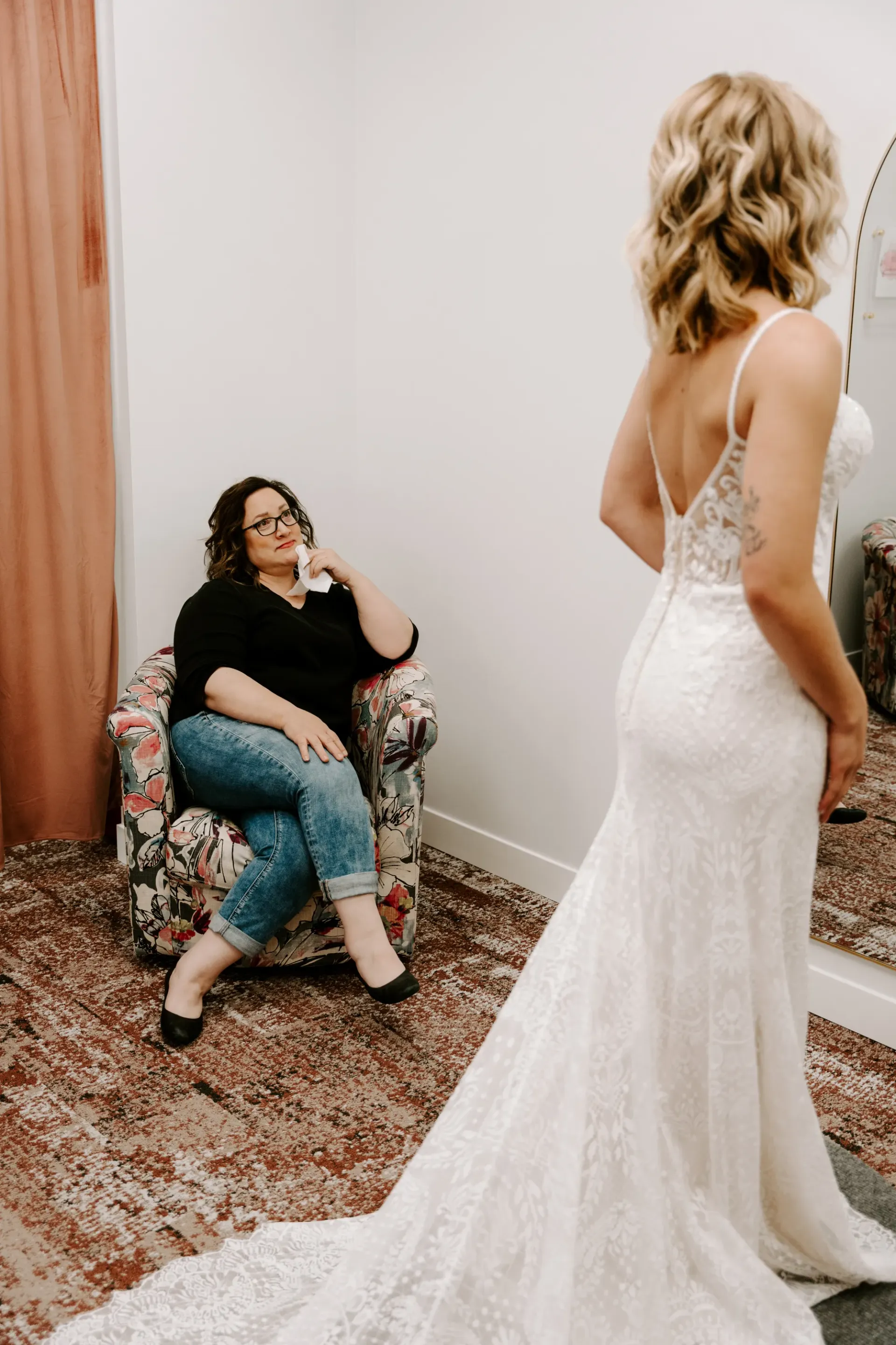 Woman in a wedding dress in a fitting room; another woman watches and smiles.