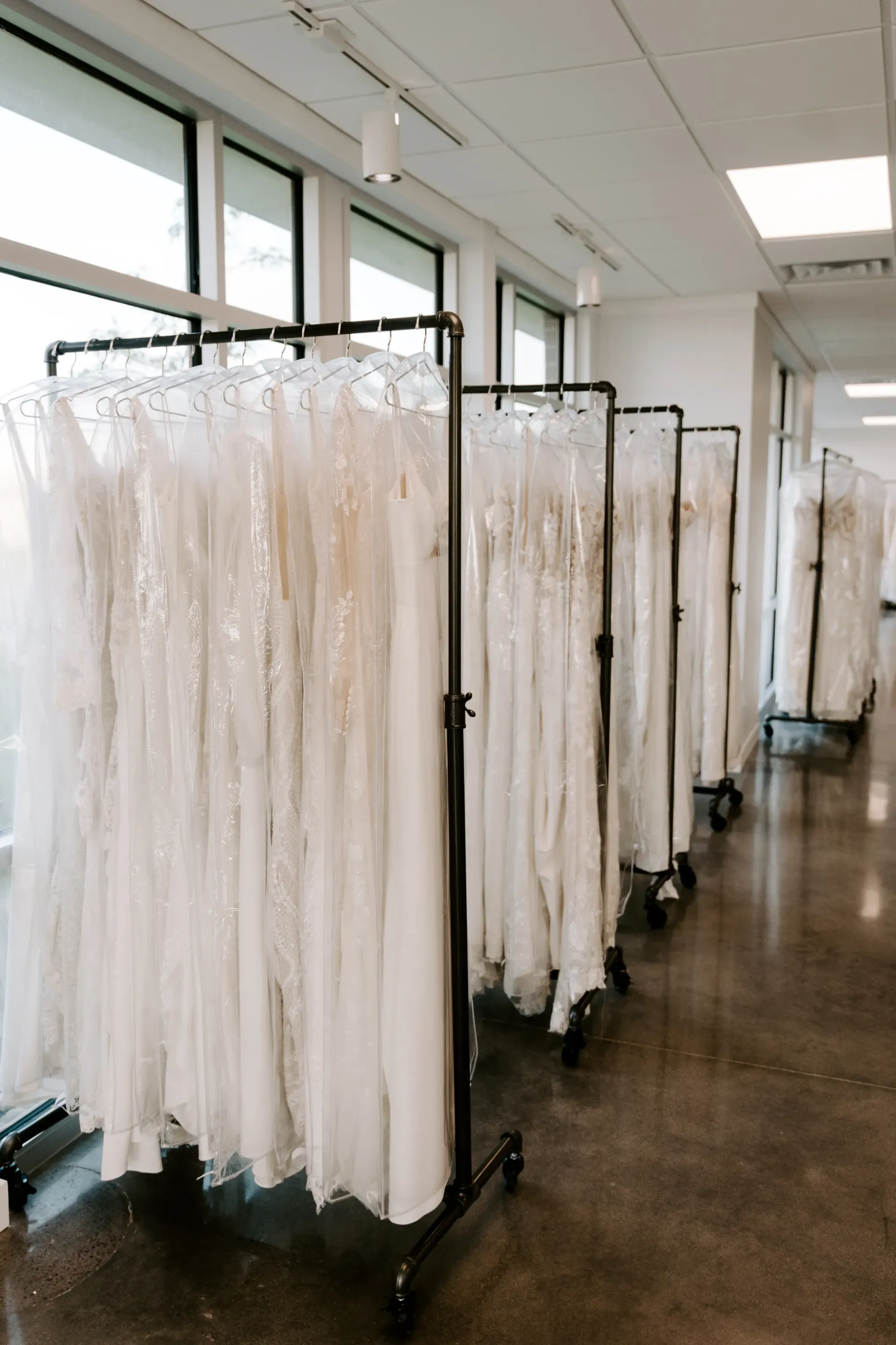 Rows of white wedding dresses in plastic garment bags on rolling racks in a bridal shop.