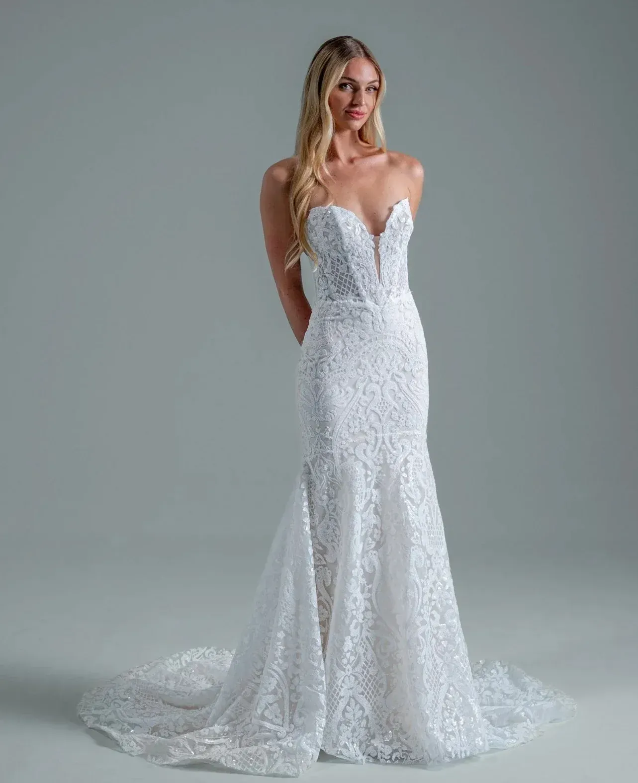 Blonde woman in a white, sparkly mermaid wedding dress with a train, poses indoors.