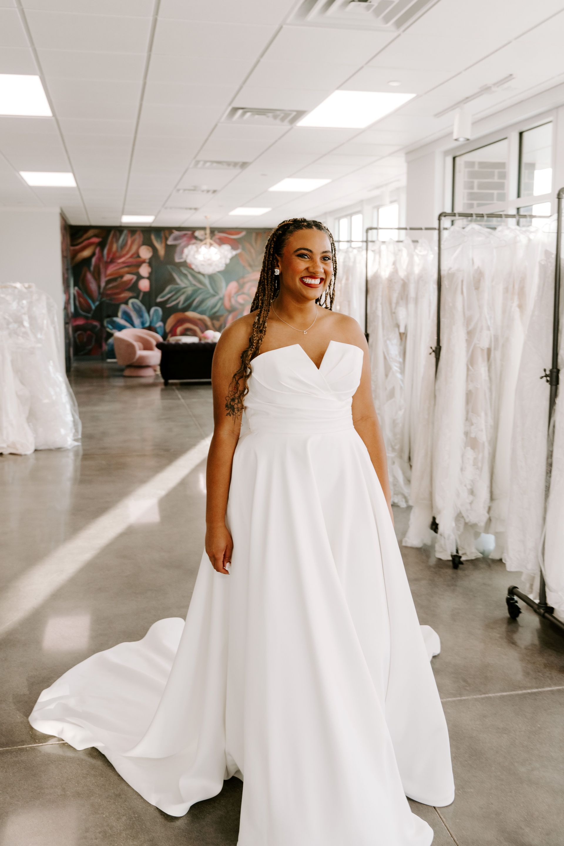 Woman in white strapless wedding dress smiles in a bridal shop with floral wallpaper and dresses on racks.
